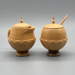 May include: A two-piece wooden serving set. The set includes a small creamer with a handle and a lidded sugar bowl with a spoon. Both pieces are light brown with a decorative scalloped edge and a pointed finial on the lid.