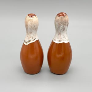 May include: A pair of ceramic salt and pepper shakers. The shakers are shaped like bowling pins. The base is a rich brown color, transitioning to a white speckled top. The top has a small hole for dispensing.