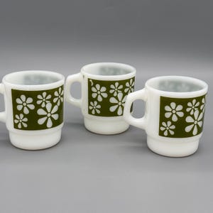 Puede incluir: Tres tazas de cerámica blanca con un diseño floral verde cuadrado. Cada taza tiene un asa y una base apilada. El diseño floral presenta flores blancas en forma de margarita. Las tazas están sobre un fondo gris.