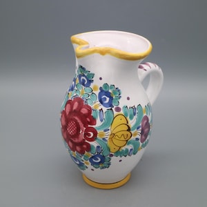 Peut inclure: Pichet en céramique blanche avec un motif floral. Le pichet présente une fleur rouge, des accents bleus et violets et un papillon jaune. Le bord et la base sont bordés de jaune. La poignée a des rayures violettes.