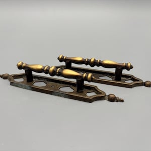 May include: Two antique brass drawer pulls. Each pull features a decorative handle with a detailed design and a rectangular base with a geometric pattern. The pulls have a vintage aesthetic and are likely for furniture restoration.