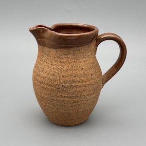 May include: A handmade, speckled stoneware jug with a brown rim and handle. The jug has a rounded body and a spout for easy pouring. The earthy tones and textured surface give it a rustic, artisanal feel. Ideal for serving beverages.