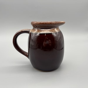 May include: A glossy, dark brown ceramic mug with a rounded body and a curved handle. The rim and top edge feature a lighter brown glaze. The mug is designed for beverages and has a classic, functional shape.
