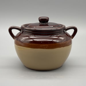 Puede incluir: Una olla de cerámica de dos tonos con tapa. La olla tiene una parte superior y tapa de color marrón oscuro, una banda marrón claro y una base beige. Cuenta con dos asas y un pequeño pomo en la tapa. La olla probablemente se utiliza para cocinar o almacenar.