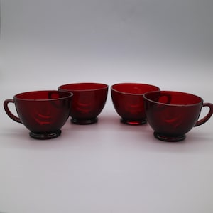 Puede incluir: Cuatro tazas de té de vidrio rojo rubí con asas. Las tazas tienen forma de cuenco redondeado y se asientan sobre una pequeña base circular. Las asas son curvas y están unidas al lateral de la taza. Las tazas están espaciadas uniformemente.