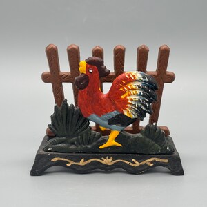 May include: A cast iron rooster figurine in red, yellow, and blue. The rooster is positioned in front of a brown fence and green foliage. The base is black with gold detailing. A decorative farm animal figurine.