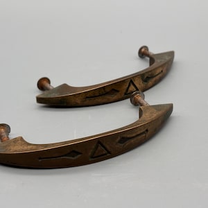 May include: Two antique bronze-colored cabinet handles. Each handle has a curved design with a decorative triangle and diamond pattern. The handles are attached with screws.