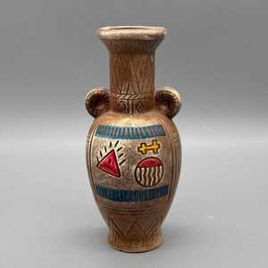 Peut inclure: Un vase en céramique marron avec une surface texturée et des poignées décoratives. Le vase présente des rayures horizontales bleues et des formes géométriques colorées, dont un triangle rouge et une croix jaune. Le vase mesure environ 15 cm de haut.