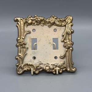May include: Ornate, gold-colored double light switch cover with a floral and scroll design. The cover has two switch openings and is made of metal. The background is a light gray.