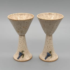 Może przedstawiać: Dwie nakrapiane ceramiczne szklanki do martini. Szklanki mają szeroką, stożkową misę na smukłej nóżce, która rozszerza się u podstawy. Kiść niebieskich winogron zdobi podstawę każdej szklanki. Szklanki są beżowe z brązowymi plamkami.