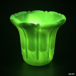 May include: A glowing neon green glass vase with a scalloped rim and vertical ribbed detailing. The translucent material emits a bright light, highlighting the curved silhouette and the textured surface against a dark, solid black background.