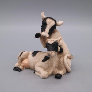 Op de afbeelding: Een beeldje van twee koeien, de ene bovenop de andere. De koeien zijn voornamelijk crèmekleurig met zwarte vlekken op hun lichaam en gezicht. Het beeldje heeft een gedetailleerd, realistisch uiterlijk, met zichtbare hoorns en hoeven.