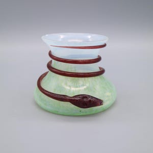 Może przedstawiać: Mały, zielony i biały szklany wazon z owiniętym wokół niego czerwonym szklanym wężem.