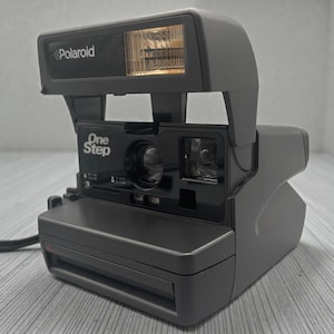 Peut inclure: Un appareil photo instantané Polaroid OneStep gris. L'appareil photo est équipé d'un flash intégré et le mot "Polaroid" est imprimé sur le dessus. Les mots "One Step" sont imprimés sur le devant. Mesure environ 15 cm de long.