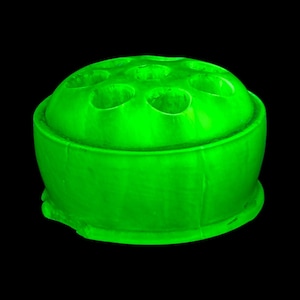 Op de afbeelding: Een felgroene bloemenkikker, een ronde container met meerdere gaten voor het rangschikken van bloemen. De kikker heeft een brede basis en een koepelvormige bovenkant met verschillende openingen. Het object staat tegen een zwarte achtergrond.