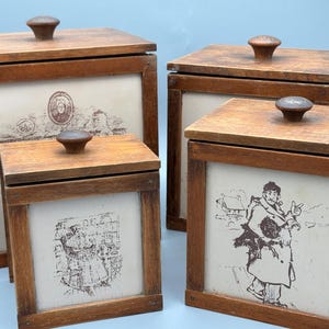 Może przedstawiać: Cztery antyczne drewniane pojemniki do przechowywania z pokrywkami i dekoracyjnymi ilustracjami. Pudełka są w różnych rozmiarach, z brązowymi drewnianymi ramami i pokrywkami. Ilustracje są w odcieniach sepii i wydają się być zabytkowymi grafikami.