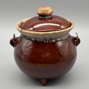 以下が含まれることがあります： 蓋付きの茶色の陶器の鍋。鍋は丸みを帯びた形で、3つの小さな足と2つの金属製のハンドルが付いています。蓋の上部にはつまみがあります。鍋の縁とつまみは明るい色で、濃い茶色の本体とのコントラストを生み出しています。