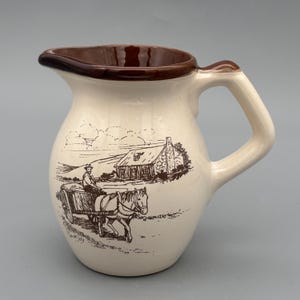 Puede incluir: Jarra de cerámica color crema con borde y asa marrones. La jarra presenta una ilustración en tonos sepia de un carro tirado por caballos y una casa de campo. El asa es curva y el pico está diseñado para verter fácilmente.