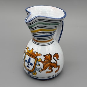 May include: White ceramic pitcher with a wavy rim and a handle. The pitcher features a blue, green, yellow, and black striped neck. A coat of arms with a fleur-de-lis and a lion is painted on the body. The pitcher is a decorative item.