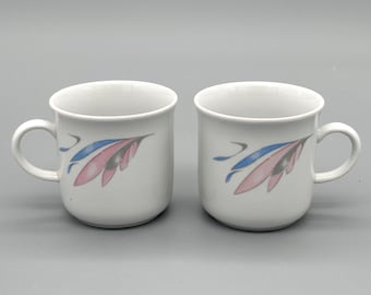 Peut inclure: Deux tasses en céramique blanche avec une anse incurvée. Chaque tasse présente un motif de feuilles roses et bleues. Les tasses sont posées sur un fond gris.