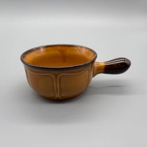 May include: A ceramic soup bowl with a handle. The bowl is a warm, earthy brown colour with a darker brown rim and handle. The interior of the bowl is a lighter shade of brown. The bowl has a classic, slightly fluted design.
