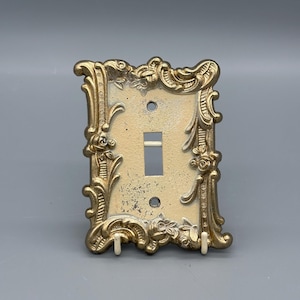 May include: Ornate, vintage-style light switch cover in gold. The cover features a rectangular opening for the switch and two screw holes. The decorative frame is embellished with floral and scrollwork details, giving it an antique aesthetic.