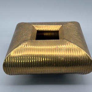 Könnte beinhalten: Ein quadratischer, messingfarbener Aschenbecher mit einer zentralen rechteckigen Öffnung. Der Aschenbecher hat eine gerippte Textur an seinen gebogenen Seiten und einen glatten, polierten Innenraum. Das Design ist geometrisch und vintage.