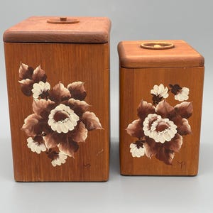 Könnte beinhalten: Zwei Holzkannen mit Deckeln. Die Kannen sind braun mit handgemalten Blumendesigns in Weiß, Braun und Creme. Die Blumen sind auf der Vorderseite jeder Kanne zentriert. Die Deckel haben kleine, runde Knöpfe.