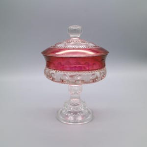 May include: A clear glass candy dish with a ruby red rim and lid. The dish has a pedestal base and a decorative, patterned design. The lid has a small, round knob. The candy dish is likely vintage or antique.