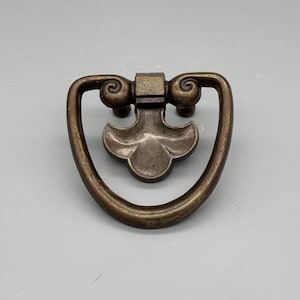 Pode incluir: Puxador de gaveta antigo em tom bronze com um design decorativo. O puxador apresenta uma alça curva com detalhes de rolagem e um acento central em forma de flor. Ideal para restauração de móveis.