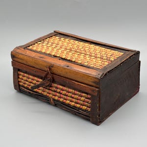 以下が含まれることがあります： 織り込まれた上部と側面を持つ長方形の木箱。上部はベージュと黄色の繊維のパターンで織られています。側面は濃い茶色の木でできており、箱は革製の留め金で固定されています。