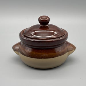 Puede incluir: Una pequeña cazuela de cerámica marrón con tapa. El plato tiene forma redondeada con dos asas laterales y una base de color beige claro. La tapa tiene un pequeño asa redondeada. El plato se utiliza probablemente para cocinar y servir.