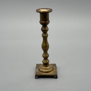 May include: A vintage brass candlestick holder with a square base and a decorative, turned stem. The candlestick is a warm, golden color and has a small, circular cup at the top to hold a candle. The overall design is classic and elegant.