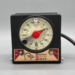 Puede incluir: Un temporizador Simmon Omega Audible Repeating Timer negro con una esfera circular de color blanco roto y números negros del 0 al 60. El dispositivo tiene una aguja central roja, un interruptor Print/Focus y un botón Start plateado.