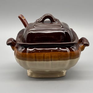 Puede incluir: Una sopera de cerámica marrón y crema con tapa y una cuchara pequeña. La sopera tiene una tapa cuadrada con asa y borde festoneado. El cuerpo de la sopera es redondo con dos asas.