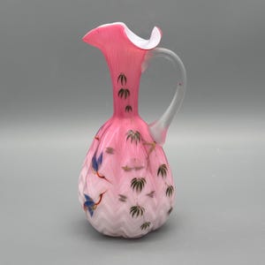 Könnte beinhalten: Rosa Glaskrug mit weißem Rand und Griff. Der Krug ist mit bemalten Kranichen und Bambusblättern verziert. Der Boden hat ein weißes und rosa Zickzackmuster.