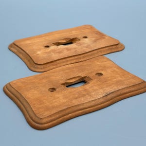 Könnte beinhalten: Zwei Holz-Lichtschalterabdeckungen mit warmbraunem Finish. Jede Abdeckung hat eine rechteckige Form mit einer geschwungenen Kante und einer einzelnen Schalteröffnung. Die Abdeckungen sind für einen Standard-Lichtschalter konzipiert.