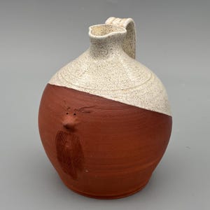 Könnte beinhalten: Handgefertigter Keramik-Krug mit einzigartigem Gesichtsdesign. Die obere Hälfte ist gesprenkelt weiß, während die untere Hälfte rotbraun ist. Der Krug hat einen Henkel und einen Ausguss. Das Gesicht hat eine Nase, Augen und einen Bart.