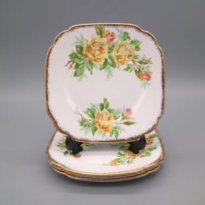 Puede incluir: Juego de cuatro platos cuadrados blancos con borde festoneado y ribete dorado. Cada plato presenta un diseño floral de rosas amarillas con hojas verdes y capullos rosas. Los platos están apilados.