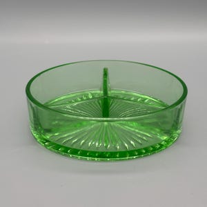 Könnte beinhalten: Eine runde, geteilte Servierschale aus grünem Glas. Die Schale ist durch einen Mittelsteg in zwei Abschnitte unterteilt. Der Boden hat ein Sonnenstrahlenmuster. Das Glas hat einen leichten Grünstich.