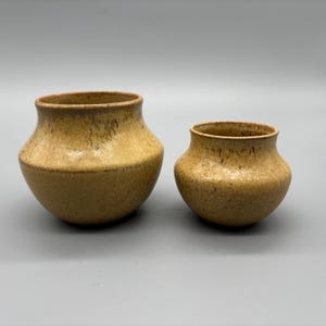 May include: Two small, handmade ceramic pots with a speckled, earthy brown glaze. The pots have a rounded shape with a wide opening and a slightly flared rim. The larger pot is approximately 13 cm in diameter, and the smaller pot is about 8 cm in diameter.