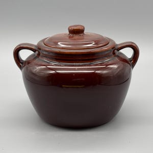 Könnte beinhalten: Ein brauner Keramiktopf mit Deckel, zwei Griffen und einer abgerundeten Form. Der Topf hat eine glänzende Oberfläche und eine dunkelbraune Farbe. Der Deckel hat einen kleinen Knopf für eine einfache Handhabung. Dieser Küchenartikel eignet sich zum Kochen und Servieren.