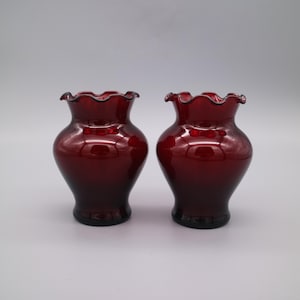 Peut inclure: Deux petits vases en verre rouge rubis avec un dessus festonné.