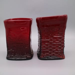 Może przedstawiać: Dwa czerwone szklane wazony z teksturowanym wzorem plecionki koszykowej. Wazony mają prostokątny kształt z lekko pochyloną górą. Szkło ma błyszczące wykończenie i głęboki czerwony kolor.