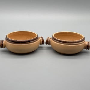 Peut inclure: Deux bols à soupe en céramique avec poignées. Les bols sont de couleur beige avec un bord et des poignées marron foncé. L'intérieur des bols est de couleur beige clair. Les bols sont posés sur un fond gris.