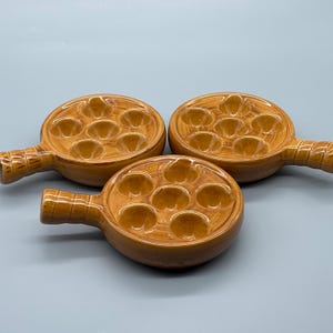 May include: Three brown ceramic escargot dishes with handles. Each dish has seven circular indentations for holding snails. The dishes are arranged on a light blue surface.
