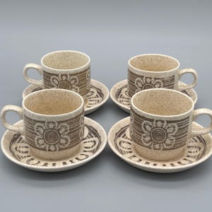 May include: A set of four vintage coffee cups and saucers. The cups and saucers are beige with a brown floral design. The cups have a handle and the saucers have a matching pattern around the edge. A classic addition to any kitchen.