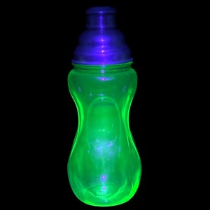 Könnte beinhalten: Eine leuchtend grüne Glasflasche mit einem dunkelblauen Deckel vor schwarzem Hintergrund. Die Flasche hat eine einzigartige Sanduhrform. Das Glas scheint von innen beleuchtet zu sein, wodurch ein lebendiger, auffälliger Effekt entsteht. Der Deckel ist ein dunklerer Blauton.