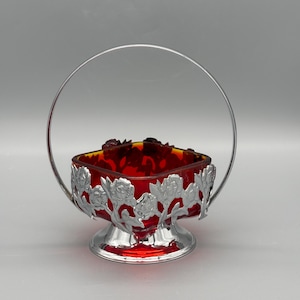 Può includere: Una bomboniera vintage in vetro rosso con un design floreale in metallo argentato e manico. Il piatto ha una forma quadrata con base arrotondata e un manico argentato che si incurva verso l'alto. Il vetro è di colore rosso intenso.