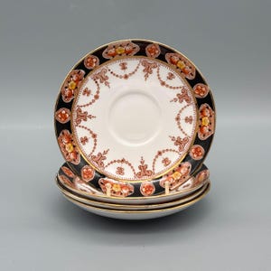 Puede incluir: Una pila de cuatro platillos antiguos. Cada platillo presenta un centro blanco, un borde negro adornado con patrones florales y geométricos en naranja, amarillo y marrón, y un borde dorado.